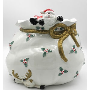 May include: A white ceramic cookie jar shaped like a sack with a gold ribbon tie. The lid features a Santa Claus figure. The sack is decorated with green holly leaves and red berries, and a reindeer figure is visible at the bottom front.
