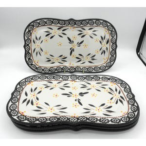 May include: Rectangular ceramic serving platters with a floral design. The white platters feature a black scalloped border and are decorated with black leaves and yellow and red flower accents. The platters are stacked on top of each other.