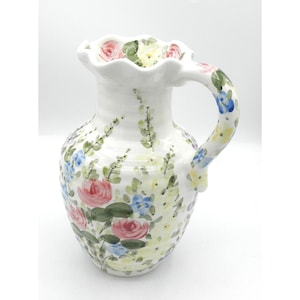 May include: White ceramic pitcher with a ruffled rim and a curved handle. The pitcher is decorated with pink roses, blue and yellow flowers, and green foliage. The floral design covers the body and handle.