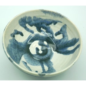 May include: A handmade ceramic bowl with a white base and blue abstract brushstrokes. The bowl has a rounded shape with a slightly raised rim. The blue design is concentrated in the center, creating a focal point.