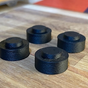 May include: Four small, black, cylindrical objects with a raised circular top are arranged on a wooden surface. The objects appear to be made of a textured material, possibly plastic or a similar substance.