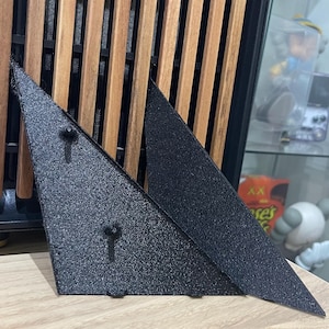 Könnte beinhalten: Zwei schwarze, dreieckige Metallteile mit strukturierter Oberfläche. Sie liegen auf einer hellen Holzoberfläche. Der Hintergrund zeigt ein Holzlattenmuster und andere Objekte.