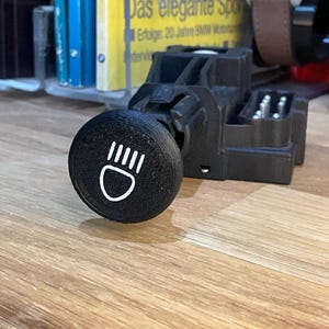 以下が含まれることがあります： 白いヘッドライトのシンボルが付いた黒いプラスチック製の車のヘッドライトスイッチ。 スイッチは、おそらく車両のダッシュボード用の、より大きな黒いプラスチックコンポーネントの一部です。 背景には雑誌と時計が含まれています。