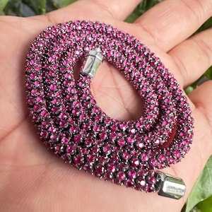 May include: A close-up of a necklace with a multitude of small, round, magenta-colored gemstones. The necklace is made of a silver-colored metal and has a clasp. The gemstones are closely set together, creating a continuous, sparkling surface.