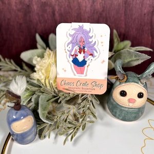May include: A magnetic bookmark featuring an anime character with pink hair, a white top, and a blue skirt. The bookmark has the text "Chaos Crate Shop" and is surrounded by greenery and ceramic figurines.