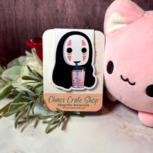 May include: A magnetic bookmark featuring a character with a white face, black hair, and a pink drink with boba. The bookmark has the text "Chaos Crate Shop" and "Magnetic Bookmark". A pink plush cat is visible in the background.