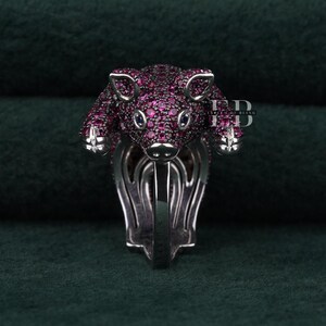 Puede incluir: Un anillo de plata con un diseño de cerdo. El cerdo está cubierto de pequeñas piedras preciosas rosas. El anillo tiene un diseño detallado y estilizado, con el cuerpo y la cabeza del cerdo prominentemente mostrados. El anillo está sobre un fondo verde oscuro.