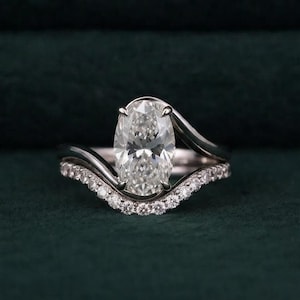 May include: A silver-toned ring featuring a large oval-cut center stone held in a prong setting. The band has a curved, asymmetrical design with a row of small, round, clear pavé-set stones along the lower edge, creating a layered appearance.