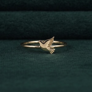 Op de afbeelding: Een goudkleurige ring met een klein, gestileerd vogelmotief in het midden van de dunne band. De vogel is afgebeeld in een vliegende positie met gespreide vleugels en heeft een gladde, gepolijste metalen afwerking over het hele sieraad.