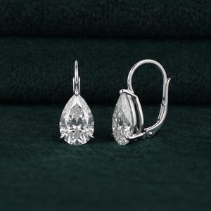 May include: A pair of elegant drop earrings featuring pear-shaped, clear gemstones set in a silver metal. The earrings have a lever-back closure and are displayed against a dark green background.
