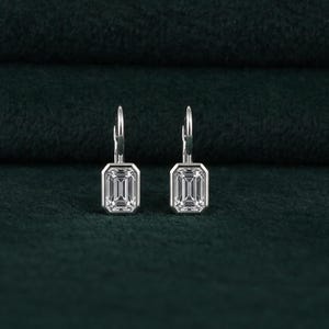 May include: A pair of silver earrings with emerald-cut, clear gemstones. The earrings have a lever-back closure and are set against a dark green background. The gemstones are rectangular with a step-cut facet pattern.