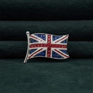May include: A silver-toned metal brooch shaped like a waving Union Jack flag. The design is filled with small, circular red, blue, and clear crystals arranged to replicate the pattern of the British flag, featuring a central cross and diagonal lines.