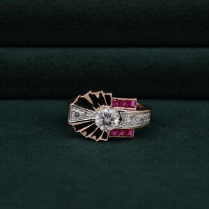 May include: A rose gold ring featuring a central round brilliant-cut diamond, flanked by black enamel fan-shaped accents, square-cut red rubies, and small round white diamonds set along the band in a geometric, multi-layered arrangement.