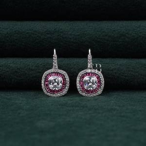May include: A pair of silver-toned earrings. Each earring features a square, clear center stone surrounded by a halo of small, pink stones. The outer edge is lined with smaller, clear stones. The earrings have a lever-back closure.