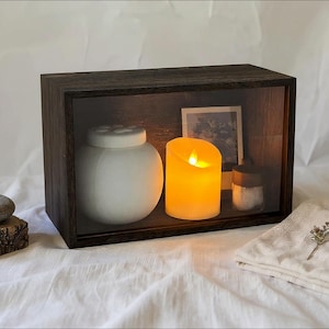 May include: A rectangular dark wood display box with a glass front containing a white spherical ceramic jar, a glowing orange LED pillar candle, a small framed photograph, and a glass jar with a dark lid, positioned near a stone and cloth.