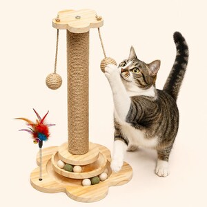 May include: A light-colored wooden cat scratching post with a flower-shaped base and top. The central post is wrapped in natural sisal rope. Two hanging sisal balls are suspended from the top, and a spring-mounted feather toy is attached to the base.