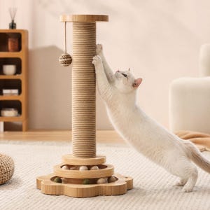 May include: A light-colored cat scratching post with a tall, rope-wrapped central column, a flower-shaped wooden base, and a circular track containing small wooden balls. A dangling rope ball toy hangs from the top of the post.