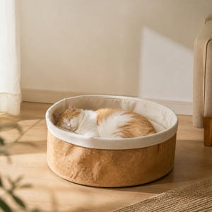 May include: A round, tan-colored pet bed with a textured, paper-like exterior and a soft, cream-colored interior lining. A ginger and white cat is curled up and sleeping inside the circular bed, which sits on a light-colored wooden floor.