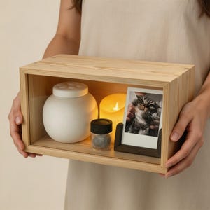 May include: A rectangular light-colored wooden shadow box display case containing a white spherical ceramic jar with a lid, a small cylindrical glass jar with a black lid, a yellow LED candle, and a framed photograph of a cat.