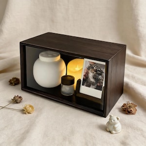 Può includere: Una scatola espositiva in legno marrone scuro con frontale in vetro, contenente un vaso bianco, un piccolo contenitore di vetro e una candela accesa. Viene visualizzata una foto polaroid di un gatto. Gli elementi decorativi includono un piccolo orso in ceramica e fiori secchi.