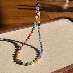 May include: A phone charm strap featuring a variety of small, round, multi-coloured glass beads. The beads display diverse patterns including floral motifs, stripes, and solid colours in shades of red, blue, green, yellow, and white. A silver-toned metal lobster clasp and a thin black cord are attached to the top.