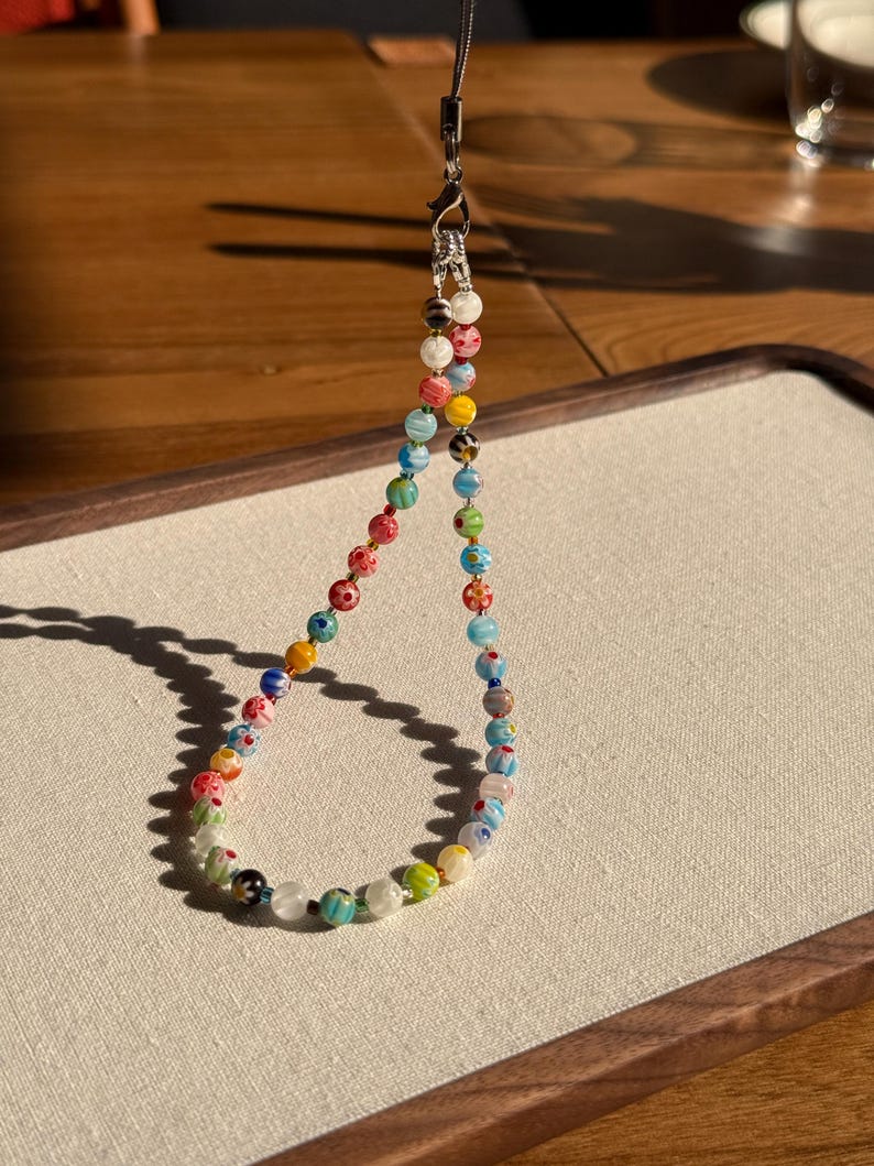May include: A phone charm strap featuring a variety of small, round, multi-coloured glass beads. The beads display diverse patterns including floral motifs, stripes, and solid colours in shades of red, blue, green, yellow, and white. A silver-toned metal lobster clasp and a thin black cord are attached to the top.