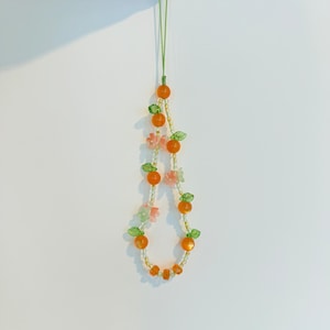 May include: A beaded phone charm featuring orange beads resembling oranges, green leaf accents, and small pink flower beads. The charm is strung on a green cord and held up against a white background.