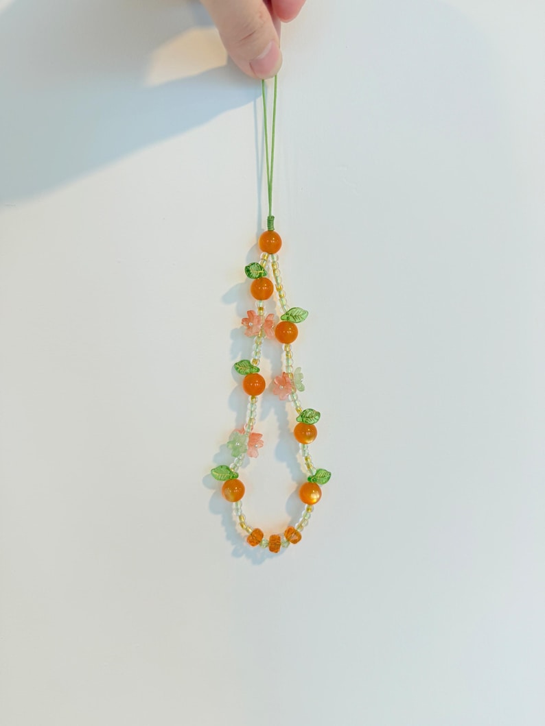 May include: A beaded phone charm featuring orange beads resembling oranges, green leaf accents, and small pink flower beads. The charm is strung on a green cord and held up against a white background.