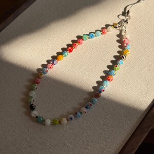May include: A phone charm strap featuring a variety of multicoloured glass beads. The beads include floral patterns, solid colours, and translucent finishes, strung together on a thin cord with a silver-toned metal lobster clasp and attachment loop.