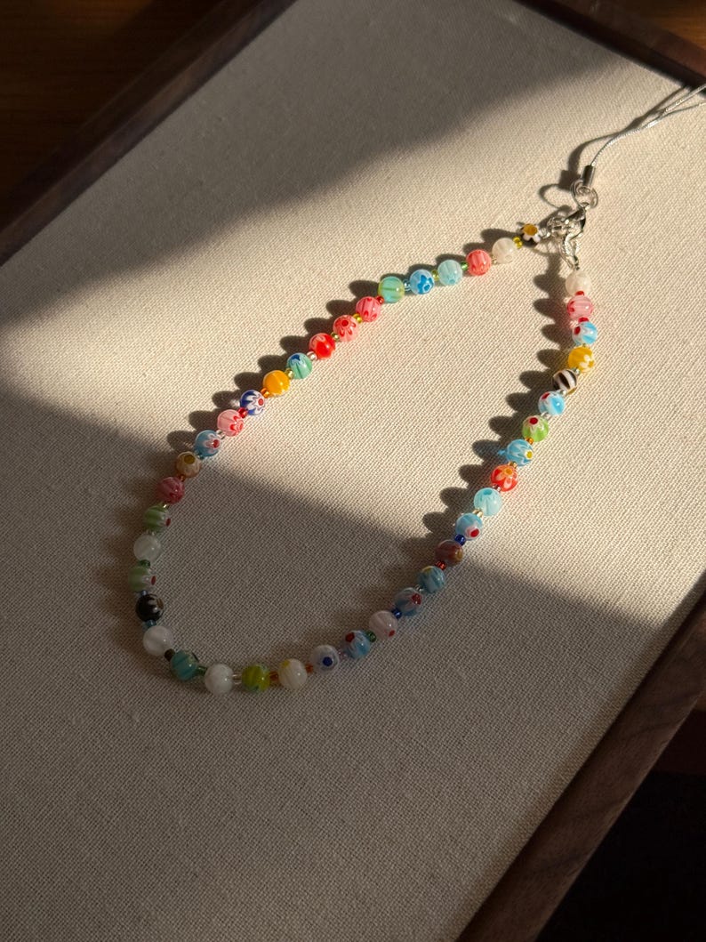 May include: A phone charm strap featuring a variety of multicoloured glass beads. The beads include floral patterns, solid colours, and translucent finishes, strung together on a thin cord with a silver-toned metal lobster clasp and attachment loop.