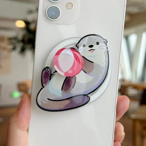 May include: A white smartphone with a pop-up grip featuring a cartoon otter holding a red and white striped ball. The otter is gray, white, and purple. The phone has two camera lenses.