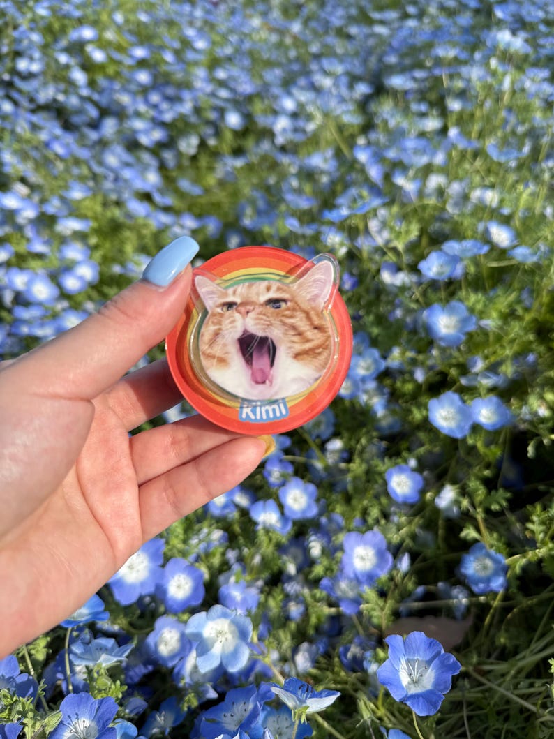 May include: A round, red cat-themed phone grip with a rainbow border. The image features an orange tabby cat with its mouth open, and the name "Kimi" is printed below the cat. The background is a field of blue flowers.