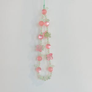 May include: A beaded phone charm featuring pink and green beads, with pink flower and fruit-shaped accents. The charm is suspended by a thin, light green string, creating a delicate and whimsical accessory.