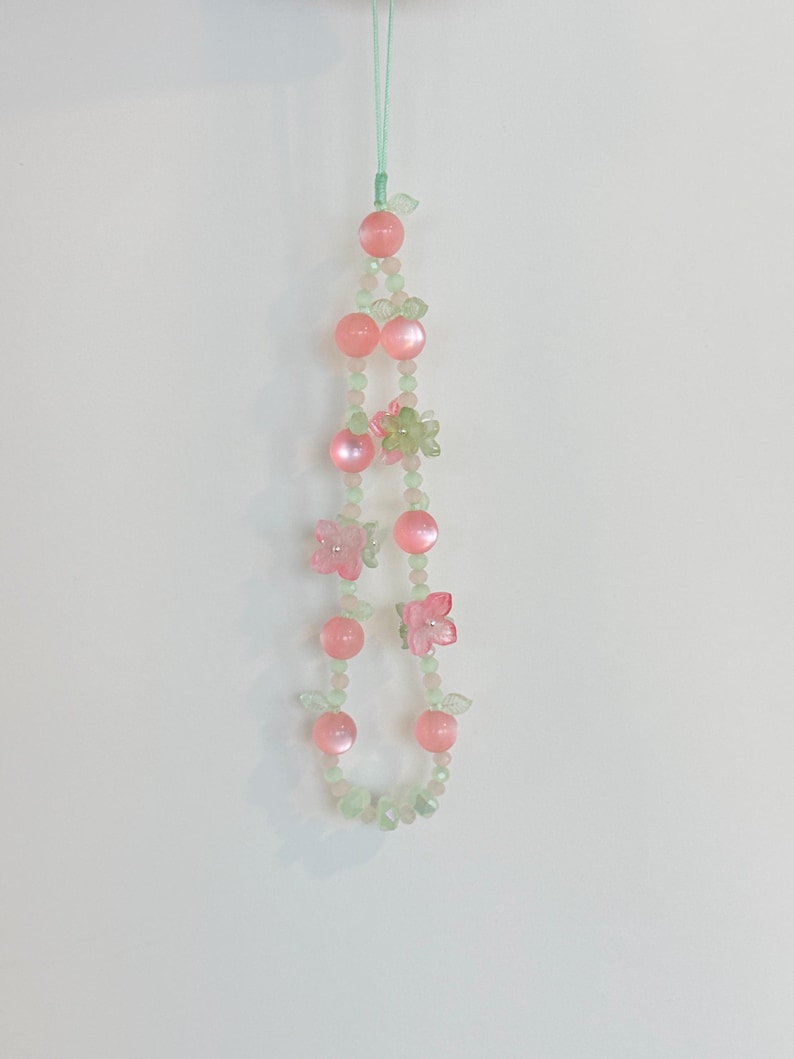 May include: A beaded phone charm featuring pink and green beads, with pink flower and fruit-shaped accents. The charm is suspended by a thin, light green string, creating a delicate and whimsical accessory.