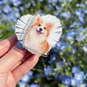 May include: A heart-shaped acrylic charm featuring a smiling corgi dog illustration. The charm has a white background with radial lines and a small hole for attachment. The corgi has orange and white fur, with a pink tongue. The background is a field of blue flowers.
