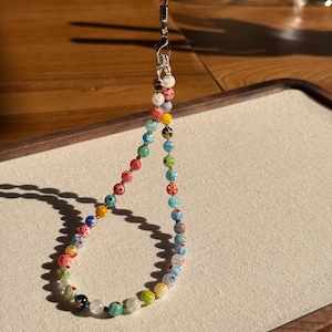 May include: A multi-coloured beaded phone strap with a silver clasp. The beads feature a variety of patterns and colours, including red, blue, yellow, and green. The strap is displayed against a neutral background.