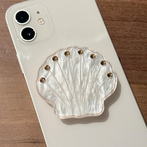 Könnte beinhalten: Ein weißer Muschel-Telefonhalter mit goldfarbenen Verzierungen ist auf der Rückseite eines weißen Smartphones befestigt. Das Muscheldesign hat ein perlmuttartiges Finish und ist fächerförmig.