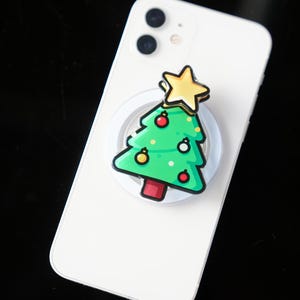May include: A white smartphone with a Christmas tree phone grip attached. The green tree has red and yellow ornaments and a gold star on top. The phone grip is round and white.