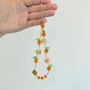 May include: A beaded phone charm featuring orange-colored beads resembling oranges, interspersed with green leaves and pink flower beads. The charm is held by a green cord.
