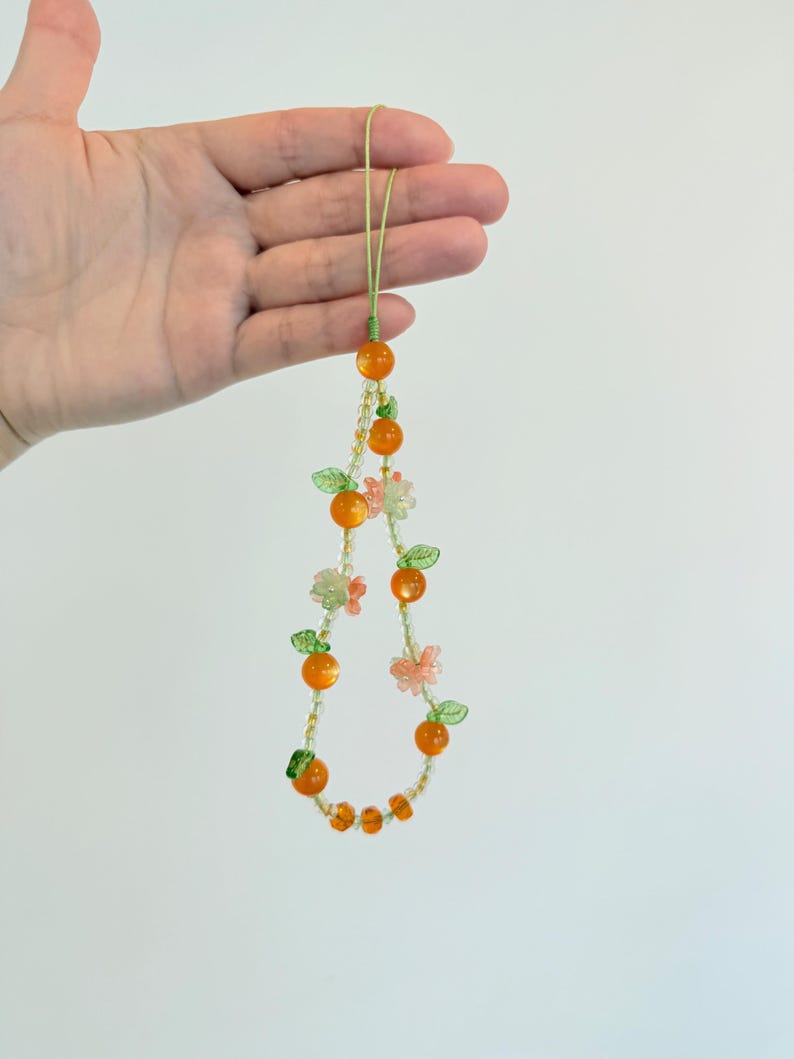 May include: A beaded phone charm featuring orange-colored beads resembling oranges, interspersed with green leaves and pink flower beads. The charm is held by a green cord.