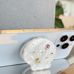 May include: A white smartphone with a gold frame and a seashell-shaped phone grip. The grip is white with a pearlized finish and adorned with various colorful gemstones. The phone is resting on a reflective surface.