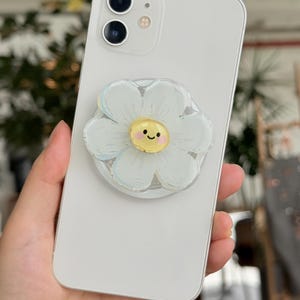May include: A white smartphone with a daisy-shaped phone grip attached. The phone grip features a white flower with a yellow center and a smiling face. The phone is held in a hand.