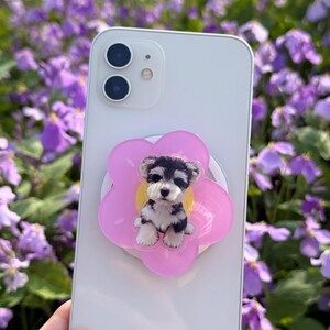 May include: A white smartphone with a pink flower-shaped phone grip featuring a black and white puppy illustration. The phone has a dual-lens camera and is held against a backdrop of purple flowers.