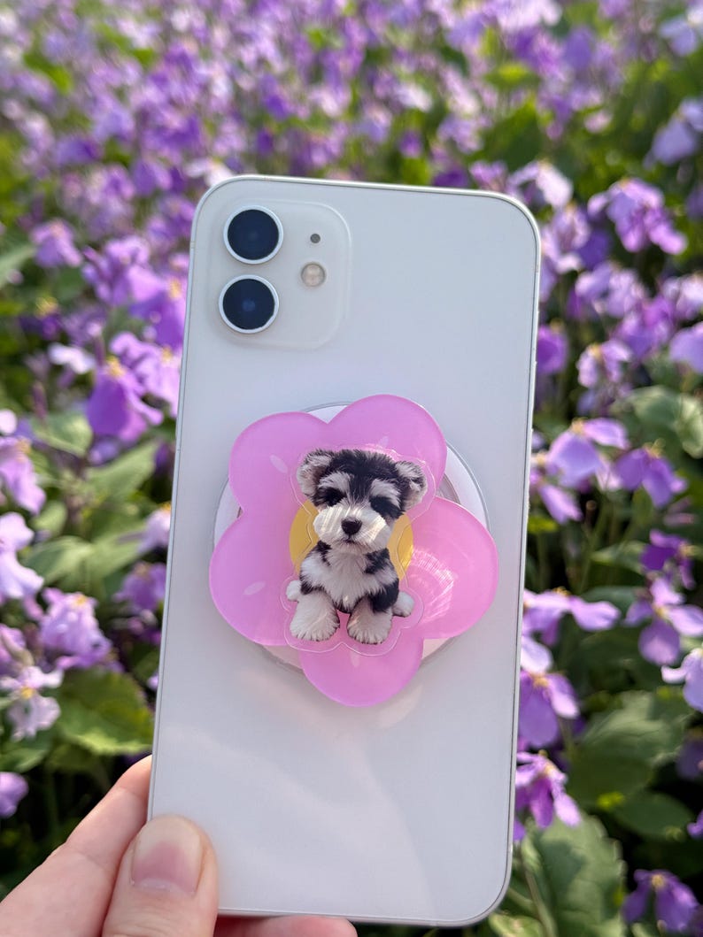 May include: A white smartphone with a pink flower-shaped phone grip featuring a black and white puppy illustration. The phone has a dual-lens camera and is held against a backdrop of purple flowers.