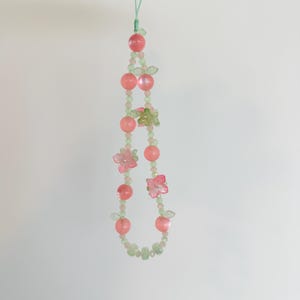 May include: A delicate beaded phone charm featuring pink and green beads, small pink flower accents, and a light green cord. The charm is strung with a variety of bead shapes and sizes, creating a whimsical and feminine design.