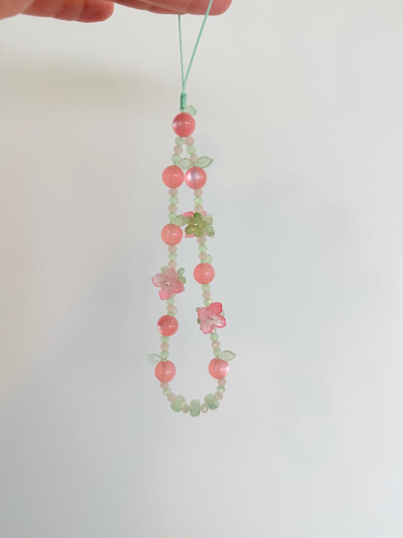 May include: A delicate beaded phone charm featuring pink and green beads, small pink flower accents, and a light green cord. The charm is strung with a variety of bead shapes and sizes, creating a whimsical and feminine design.