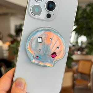 May include: A pearlescent seashell phone grip with iridescent highlights and embedded gemstones in pink, clear, and red. The grip is attached to a silver smartphone, offering a decorative and functional accessory.