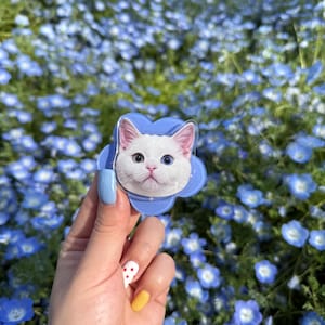 May include: A white cat with blue eyes is featured on a blue flower-shaped phone charm. The charm is held by a hand with blue and white polka dot nail polish.