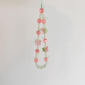 May include: A beaded phone charm featuring pink and green beads, with pink spheres and small flower and butterfly accents. The charm is suspended from a light green cord.