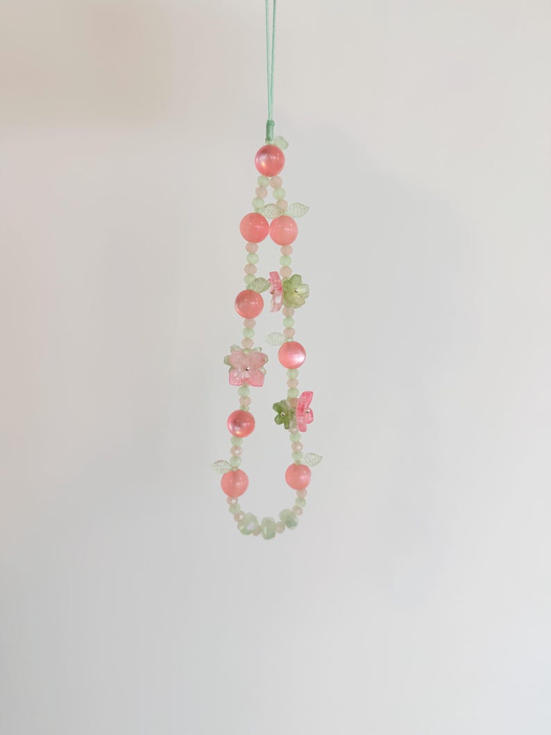 May include: A beaded phone charm featuring pink and green beads, with pink spheres and small flower and butterfly accents. The charm is suspended from a light green cord.